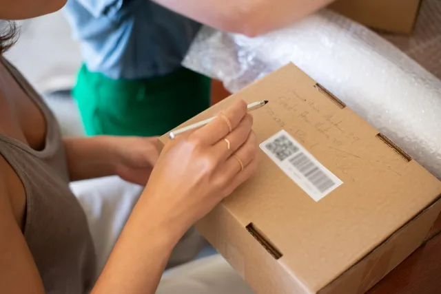 Close-up of a woman writing on a cardboard box, preparing for shipping. Jak zorganizować zwroty w e-commerce: proces od zgłoszenia do refundacji