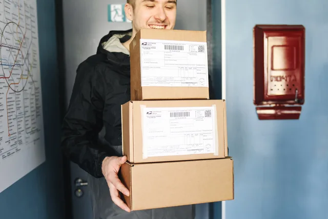 Smiling delivery man holding packages inside a building, ready for delivery. Koszty wysyłki: jak ułożyć cennik i darmową dostawę?
